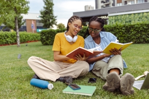 Two young female students reading books together and cheerfully discussing, while sitting on a grass area
