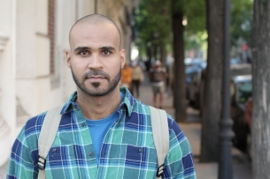 A man with a short haircut, goatee and plaid shirt, wearing a backpack, walks on a city sidewalk.