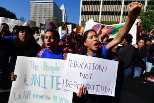 Two young people yell while holding signs, one raising a fist. A sign reads "education not deportation."