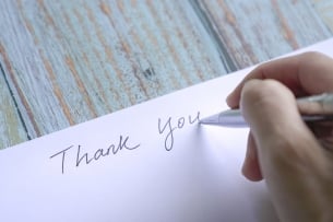 A close-up image of the hand of a person holding a pen as they write "Thank you" on a blank note card.