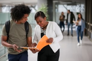 Happy teacher talking to an African American college student in the hall at the school and pointing at his notebook