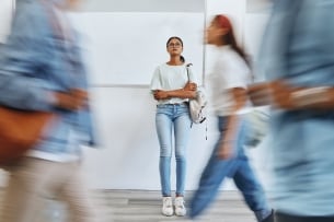 Young woman stands still in a hallway looking pensive as other young people rush by.