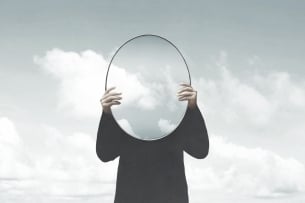An illustration of a woman holding an oval mirror up to her face against a cloudy gray-sky background.