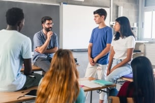 A male instructor engages in discussion with a small group of five students variously standing/sitting in a circle.