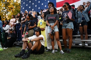 People sit and stand on a patch of grass in front of an American flag