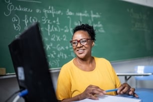 Middle aged woman with dark skin and dark hair and glasses sits in front of a computer screen with a chalkboard with math problems behind her.