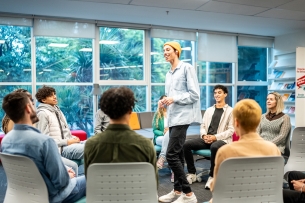 Young man in casual clothes stands in the middle of a seated circle of peers and a facilitator.