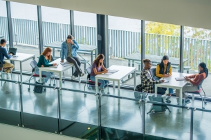 University students are seen scattered at various desks in an open study area, as they work on assignments between classes. Some students are working together while others are seen working independently in this aerial view.