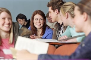 In the forefront of the picture, group of five students are gathered in a lecture hall, talking and smiling with one another.