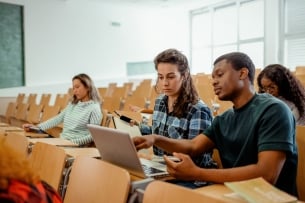Two students compare notes in a classroom with a few students sitting in the rows behind them.