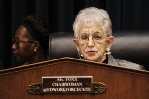 An older light-skinned woman with white hair, wearing glasses, at lectern