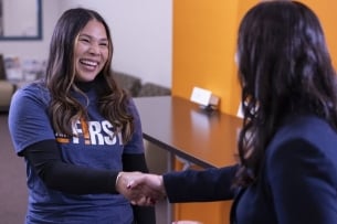 A young woman shakes another person's hand while smiling, wearing a sweatshirt that says I Am First