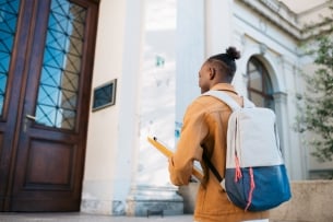 View from behind of an adult university student climbing up the steps and entering the educational building while wearing a backpack and holding a notebook