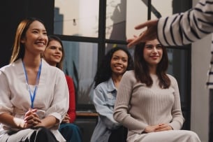 A group of four young women smile and listen attentively in the audience for a lecturer or seminar presenter.