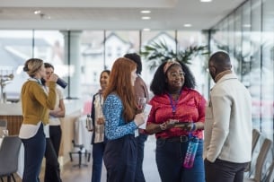A diverse group of young professionals stand chatting during a networking event; some are carrying water bottles and some are wearing lanyards with name tags.