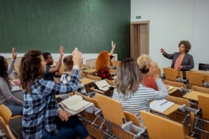 A diverse group of college students raise their hands as the instructor appears to call on one of them.