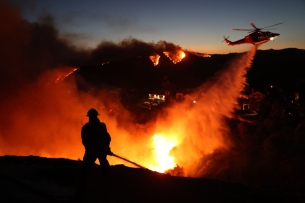 A firefighter battles a fire while a helicopter drops water on the blaze