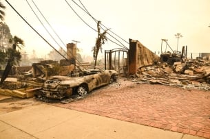 A burned out car sits in front of the ruins of a house.