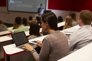 A photo of a college lecture hall taken from the back of the room -- a female student working on her laptop is in the foreground of the photo while the lecturer, presenting with slides, is visible in the background.