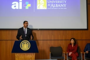 John B. King speaks at a podium, with a sign behind him reading "AI+" and "University at Albany." To his left is Governor Kathy Hochul, in a red suit.