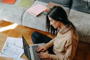 A young woman types on a laptop with papers strewn around her.