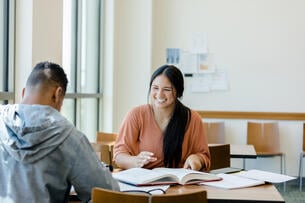 A youn woman smiles at her friend as he tries to explain the writing topic to her
