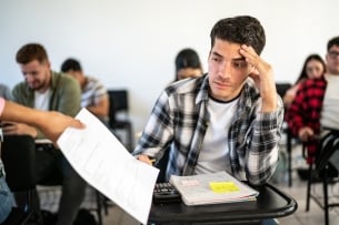 A young man with his fingers on his forehead prepares to take a graded assignment back from a professor.