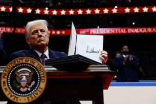 Donald Trump sits at a desk with the presidential seal, holding up an executive order he signed.