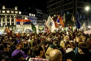 An image of a dense crowd of student protesters, some waving flags, at nighttime against the Belgrade cityscape.
