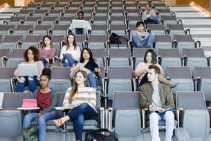 A racially diverse group of male and female college students sit in a partially filled lecture hall, with many more seats open than not. 