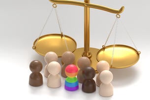 A photo depicting a golden scale, representing the scales of justice, along with nine multi-colored miniature people figurines, ranging in color from cream to tan to black, along with one that is rainbow-colored, suggesting the concept of diversity.