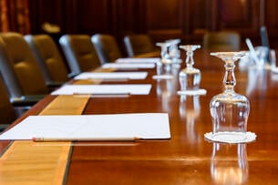 A photo of a wooden boardroom table, set up for a meeting, with pads of paper and water glasses. 