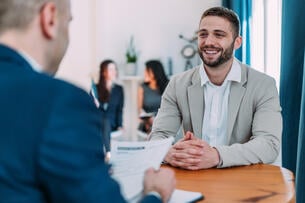 Shot of male HR manager looking through résumé and asking young man questions about his skills and previous job experience.