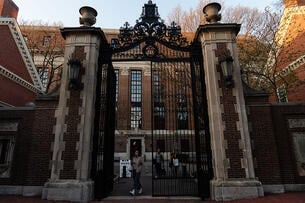 A photo of a student walking through campus gates at Harvard University, a stately brick campus building in the background.