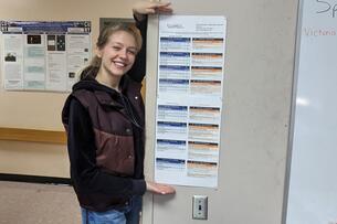 A young woman with blond hair holds up a white document with blue and orange boxes in a classroom.