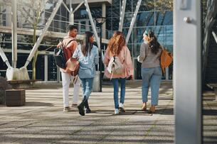 A group of four college students, each wearing backpacks, talk with one another while walking across campus.