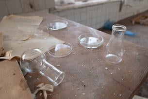 A photo of a dusty wooden table in an unused science laboratory; glass flasks, one knocked on its side, Petri dishes, and documents are strewn about the table.