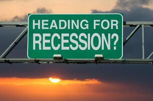A green and white road sign, as if above a highway, says "Heading for Recession?" against the backdrop of a sunset in a cloudy sky.