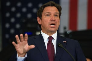 A photo of Florida Governor Ron DeSantis, a white man with brown hair, wearing a dark suit with a maroon tie and gesturing with his right hand.