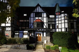 A large Tudor-style house with a red awning and trimmed hedges out front, photographed at night