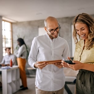 A bald man with glasses and a blonde woman with long hair stand together smiling while looking down at a tablet and smart phone.