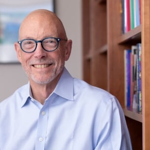 Ted Mitchell, a light-skinned man with a bald head who is wearing blue-framed glasses and a blue button-down shirt, sitting next to a bookcase