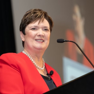 Lynn Pasquerella, a light-skinned woman with short brown hair, in a red jacket and pearls, stands at a podium