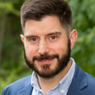 Brendan Cantwell, a light-skinned man with short dark hair and a beard and mustache, wearing a blue collared shirt under a blue jacket.