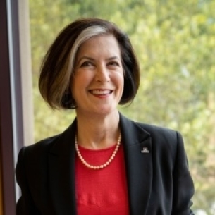 Hollis Robbins, a light-skinned woman with dark hair in a bob that has a streak of white framing her face. She is wearing a black jacket and red dress with a pearl necklace.