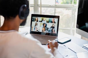 Young man looks at a computer screen with friendly-looking classmates in a grid. He is wearing headphones and faced away from the camera. 