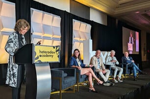 A photo of one person standing at a podium with the Heterodox Academy logo on it, to the left of four seated panelists.