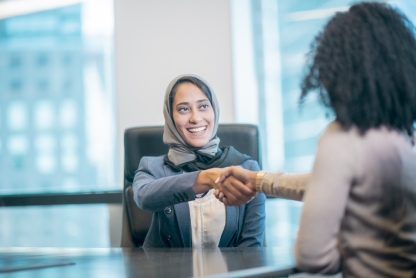 Young woman wearing headscarf shakes hands with another woman as if in an interview