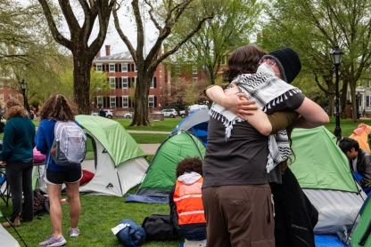 Two students, one wearing a keffiyeh, embrace in front of pitched tents at Brown University.