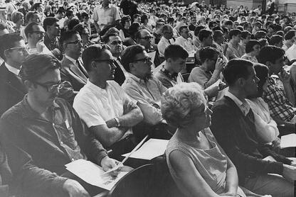 In this historical black-and-white photo from a 1965 anti-war teach-in, a dense crowd of people in an auditorium sit in an audience, seemingly listening to a speaker with intense interest and focus.